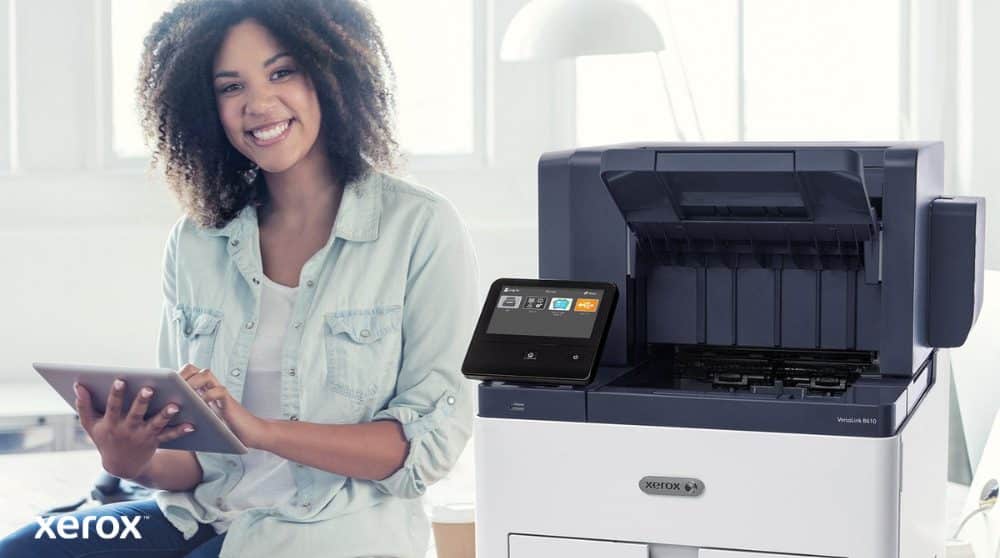 Une personne souriante est assise à côté d'une imprimante Xerox VersaLink B610 blanche, tenant une tablette et regardant la caméra, soulignant la nature conviviale des solutions Xerox.