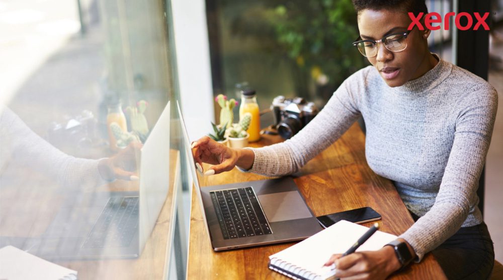 Une personne portant des lunettes travaille à une table en bois, regarde un écran d'ordinateur portable et écrit dans un cahier, avec un smartphone à proximité, suggérant un environnement de travail productif